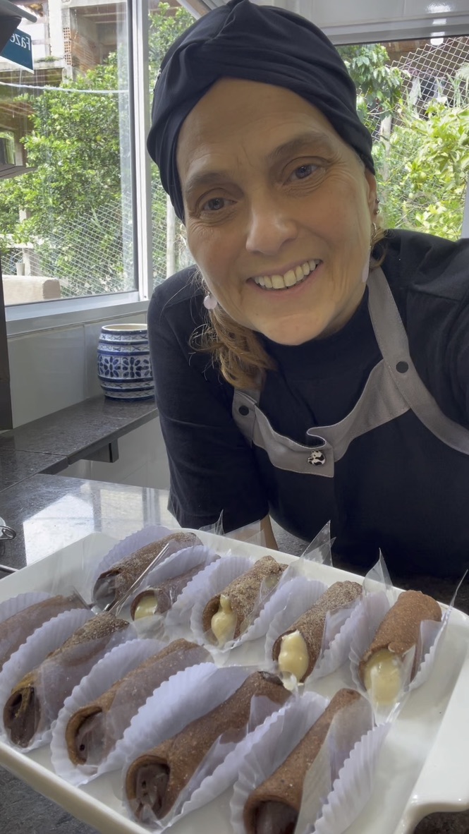 Beth Almeida em seu ateliê de panificação, sorrindo com uma bandeja de pães recém-assados